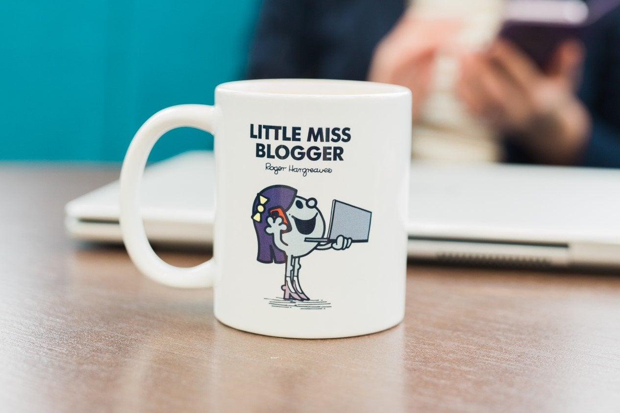 White mug with 'Little Miss Blogger' printed on it. Blurred out image of me (Eb Gargano) on my phone in the background as well as my laptop