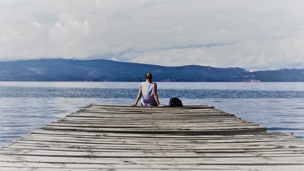 Woman sitting on a pier looking out across a lake towards hills. 