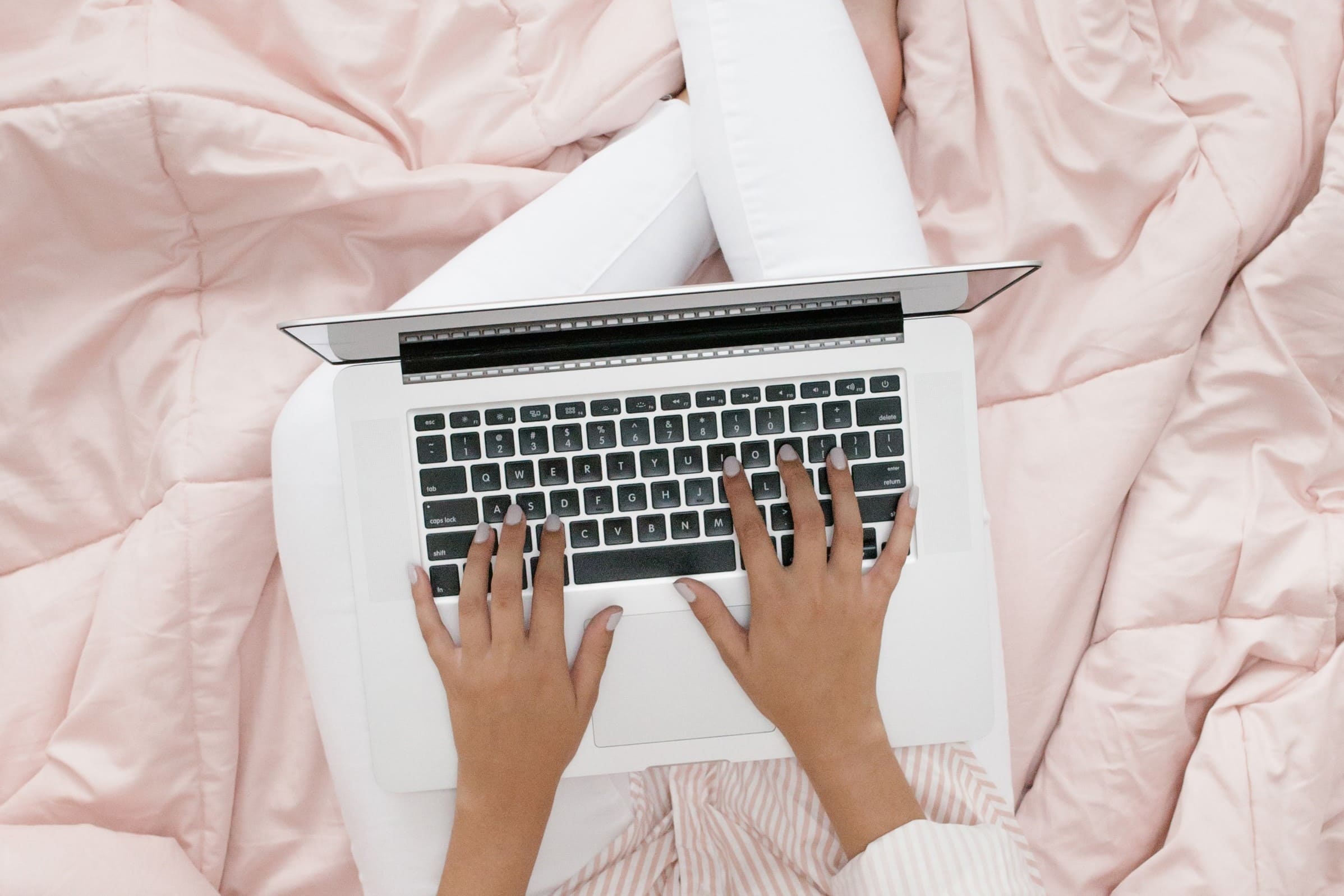 Overhead of woman typing on laptop while sitting on pink duvet